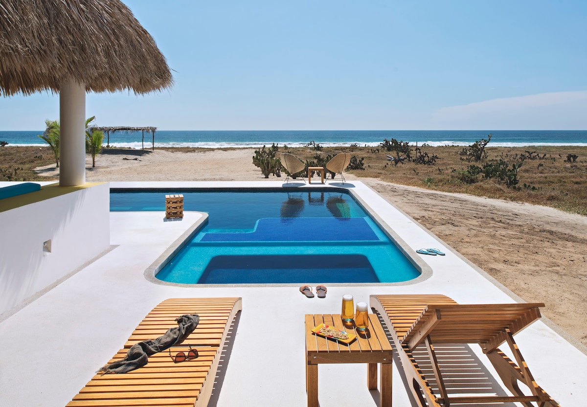 A private pool area features wooden lounge chairs beside the water, with a tropical palapa providing shade. The serene beach is visible in the background, surrounded by open land and natural vegetation. Clear blue skies enhance the inviting atmosphere of the space.