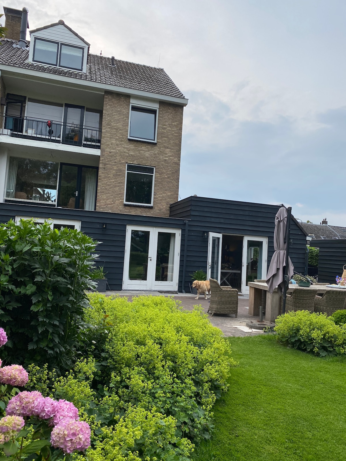 The image displays a modern family house featuring a combination of brick and dark siding. Large glass doors open to a patio area surrounded by lush greenery and blooming flowers. A shaded seating area is visible, alongside a well-maintained lawn providing a welcoming outdoor space.