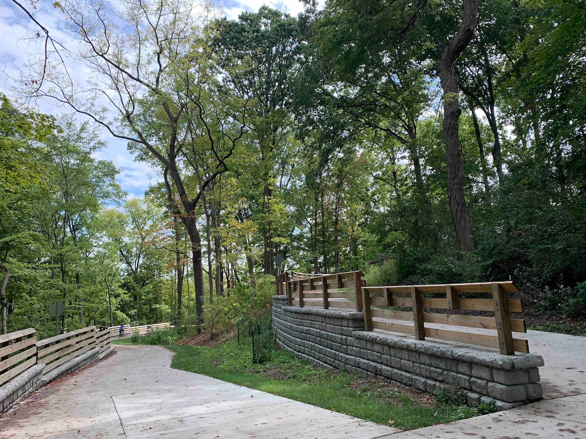 A scenic pathway winds through a lush green landscape, bordered by trees with vibrant foliage. A stone wall borders the walkway, while wooden railings provide a sense of safety along the elevated path. The trail invites leisurely walking and cycling.