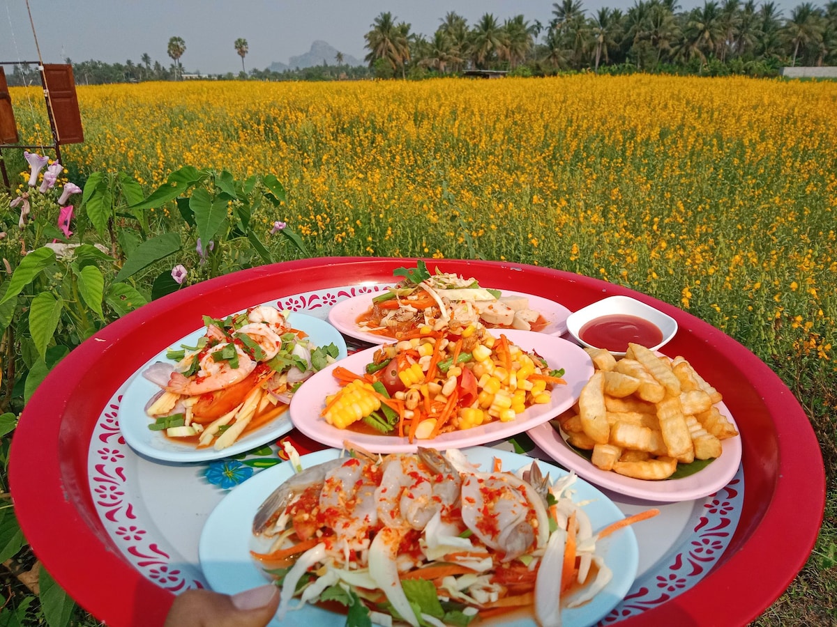 A selection of local dishes is displayed on a vibrant red tray, featuring various plates of fresh ingredients including vegetables, seafood, and dipping sauce. In the background, a sprawling field of yellow flowers and palm trees creates a tranquil countryside setting.