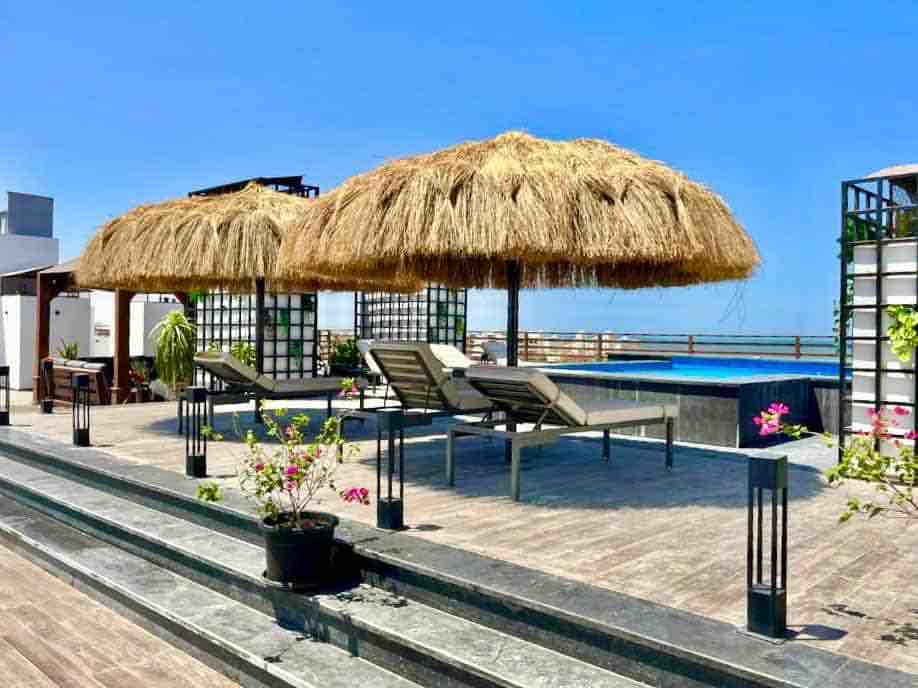 A rooftop area features two thatched umbrellas providing shade over lounge chairs. A small pool is visible in the background, surrounded by planters with blooming flowers. Hardscaped flooring and sleek lighting elements enhance the outdoor setting.