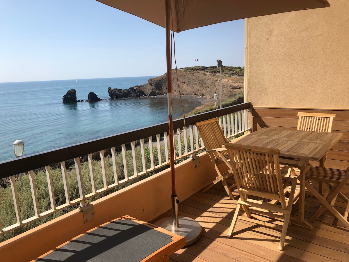 A wooden terrace features a rectangular dining table surrounded by matching chairs. A sunshade is positioned overhead, and a lounge chair is visible nearby. The ocean view includes rocky formations in the distance and calm waters, enhancing the serene atmosphere.