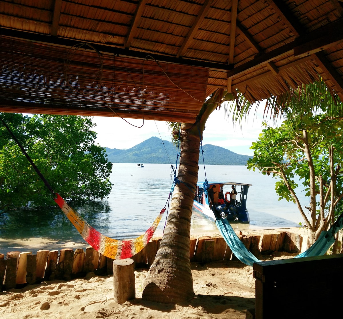 Two colorful hammocks hang between palm trees, offering a view of the calm water. A small boat approaches the sandy beach, framed by lush greenery and distant hills. The thatched roof provides shade, enhancing the serene coastal environment.