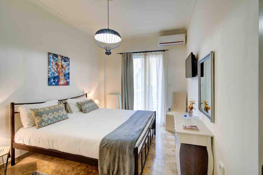 A spacious bedroom features a large bed, flanked by decorative pillows. Natural light enters through a balcony door adorned with sheer curtains. A small table with a chair and a wall-mounted television are included. An artwork is displayed above the bed, adding a thoughtful touch.