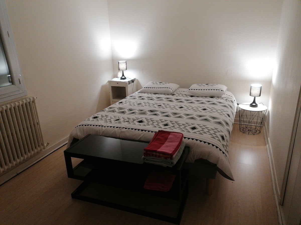 A spacious bedroom is featured with a comfortable double bed covered in a patterned blanket. Two nightstands with lamps are positioned on either side, providing soft lighting. A sleek black table is placed at the foot of the bed, displaying neatly folded towels.