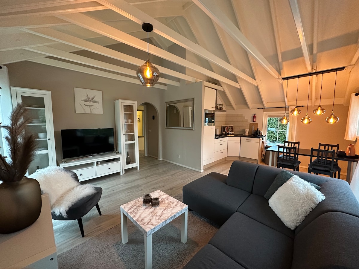 A spacious living area features a large corner sofa and a stylish marble-top coffee table. A flat-screen TV is positioned against a feature wall, while warm lighting hangs above a dining table set for six. The open kitchen is visible in the background.