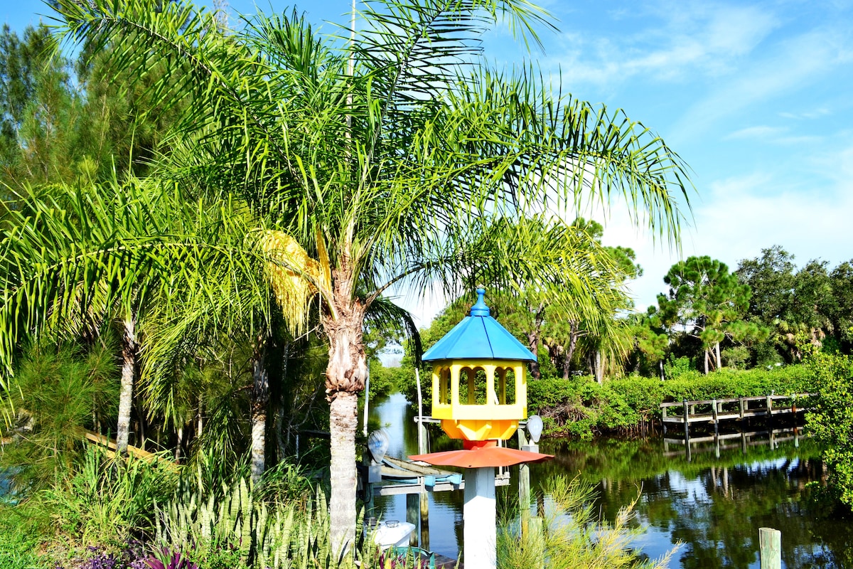 Lush palm trees frame a vibrant yellow birdhouse, situated near a serene canal. The calm water reflects the green landscape, with a glimpse of wooden docks visible in the background. A variety of native plants and textured bushes add to the natural setting.
