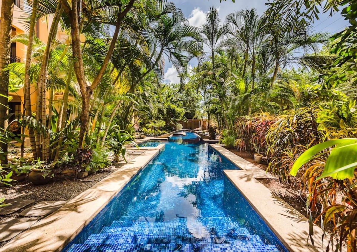 A tranquil pool area is framed by lush tropical greenery, with palm trees and vibrant foliage surrounding the water. The pool features clear, inviting blue tiles, reflecting sunlight. Pathways lined with diverse plants lead to the pool, creating a serene atmosphere for relaxation.