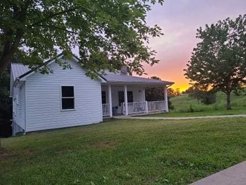 A charming white house sits on a grassy plot, framed by trees. The porch invites relaxation, and the warm glow of a sunset casts a golden hue over the surrounding landscape. The peaceful atmosphere of the countryside is highlighted by the rolling fields in the background.