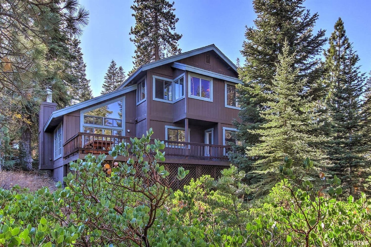 The exterior of the modern-rustic mountain house is showcased, surrounded by tall pine trees and lush greenery. A spacious deck extends from the structure, which features large windows that invite natural light while providing views of the surrounding forest.