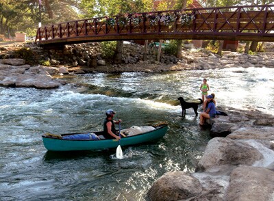 A serene river scene features a person kayaking while a dog swims nearby. Two individuals relax on rocks, enjoying the water. A wooden bridge adorned with flowers arches over the river, surrounded by greenery and natural rock formations.