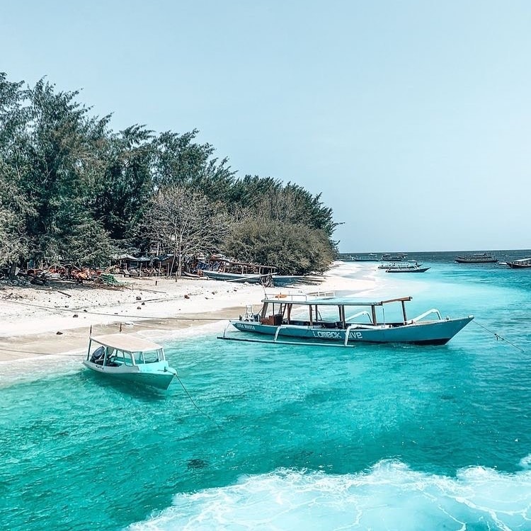 The image captures a serene beach scene on Gili Air Island, showcasing soft, white sand and clear turquoise waters. Small boats rest gently on the water, while lush greenery borders the beach, creating a tranquil atmosphere. The sky is bright and clear, enhancing the coastal views.
