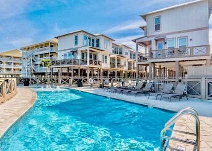 A sparkling outdoor pool is surrounded by a wooden deck and several lounge chairs. The pool area is bordered by multi-level beachfront cottages, offering a clear view of the warm sky above. The atmosphere provides a spacious area for relaxation and enjoyment.