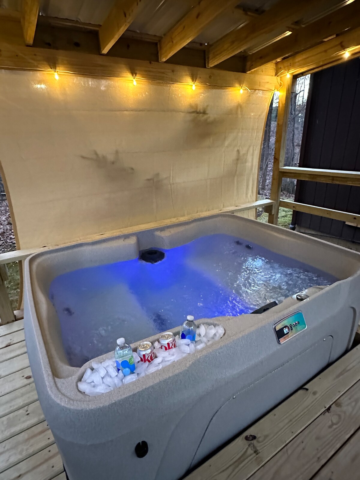 A four-person hot tub is positioned on a covered deck, featuring soft blue lighting within the water. The surrounding area is enhanced by warm string lights, creating a cozy ambiance. Refreshments are visibly arranged along the edge, inviting relaxation in a natural setting.