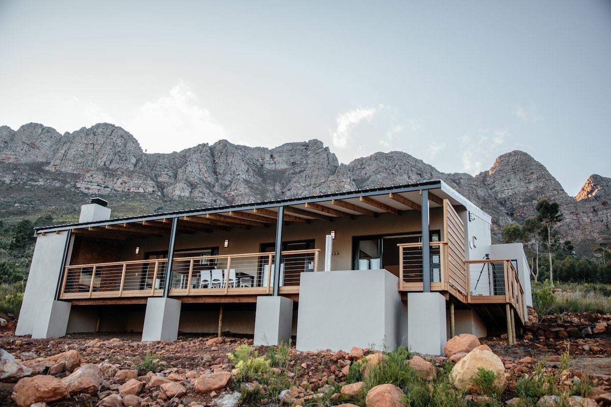 A freestanding structure is positioned among rocky terrain, with a spacious deck overlooking the landscape. Large windows are framed by wooden accents, while the backdrop features towering mountains under a clear sky, contributing to a serene atmosphere.