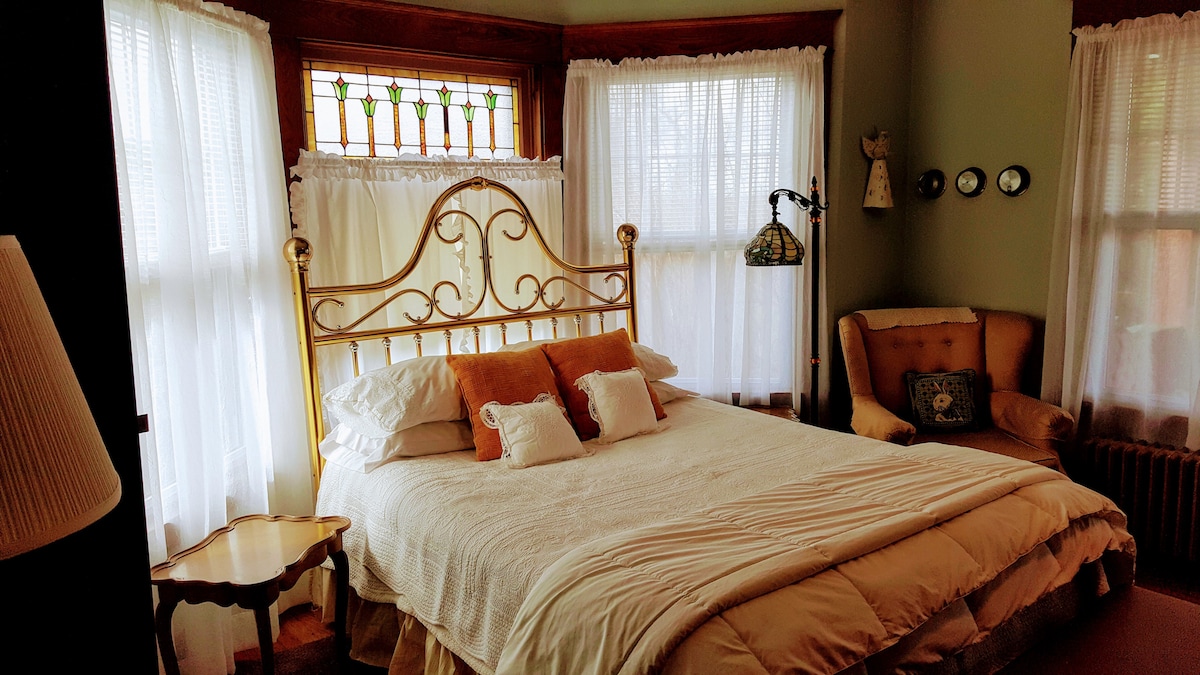 A queen-sized bed is centered in the room, adorned with crisp linens and decorative pillows. Natural light filters through sheer curtains, illuminating the stained glass window above. A cozy armchair and a small side table accompany the bed, enhancing the welcoming ambiance.