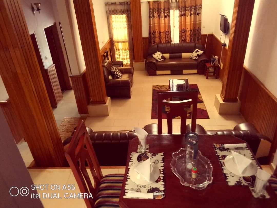 A view of the living area and dining space is provided, showcasing dark leather sofas arranged around a coffee table. A dining table is set with dishware, and decorative curtains frame the windows, allowing natural light to fill the room.