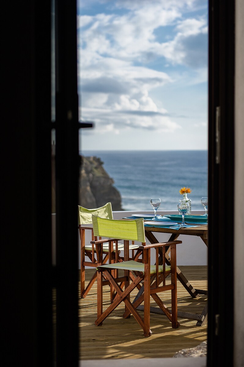 A dining area is framed by an open door, featuring a round table set for four, with glasses and a small flower centerpiece. The ocean view is visible in the background, complemented by a clear blue sky and soft clouds.