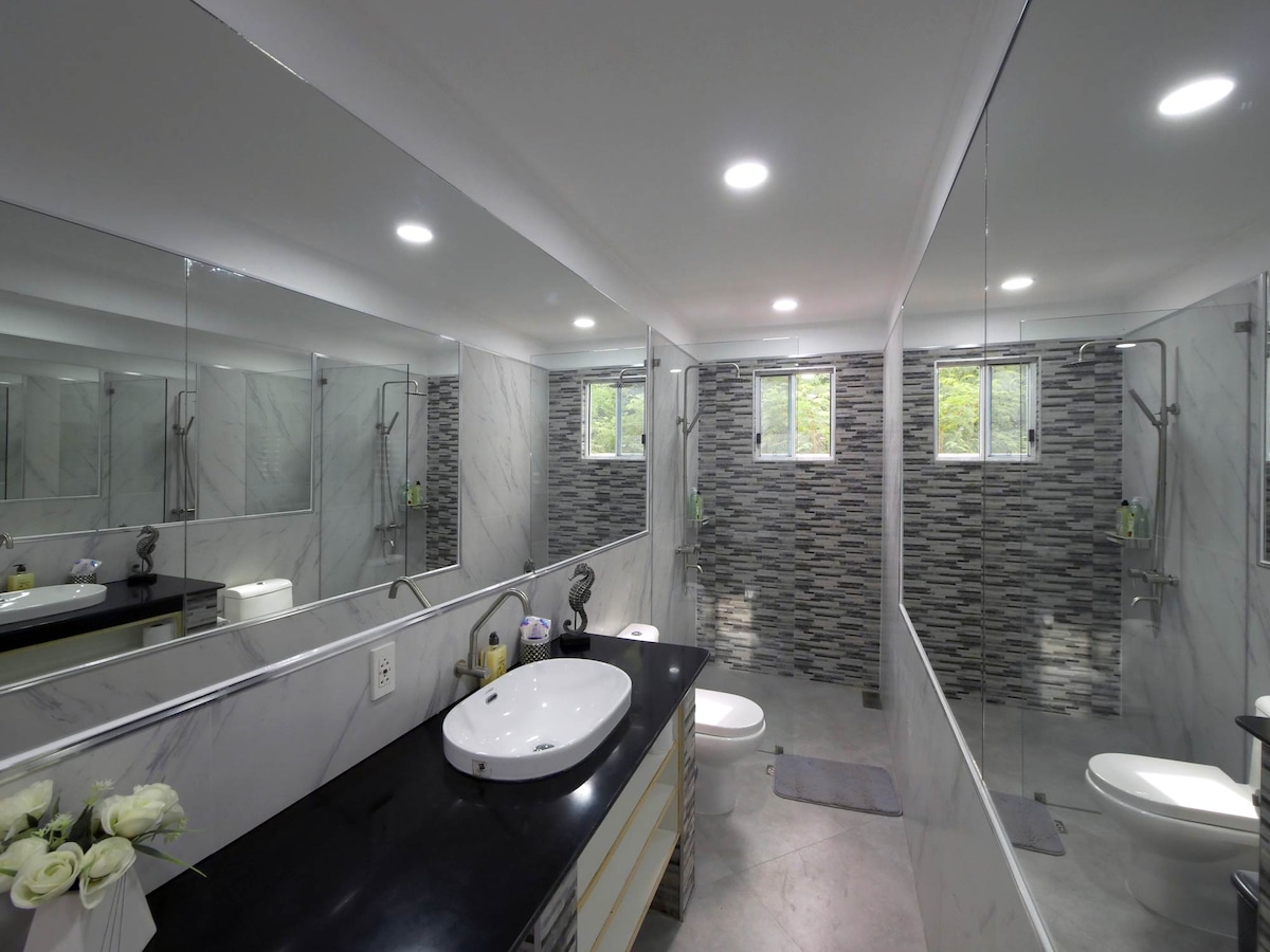 A spacious bathroom features a large dark countertop with an oval sink and modern fixtures. Full-length mirrors on opposite walls amplify the natural light from two windows. A sleek tiled shower area is adjacent to white toilet fixtures, complemented by neutral-colored tiles throughout.