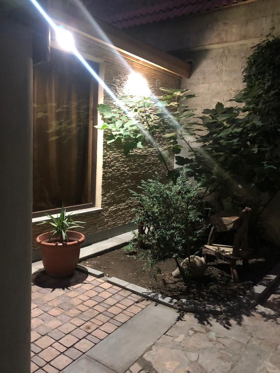 An intimate courtyard is highlighted by soft lighting illuminating the stone pathway and surrounding greenery. A potted plant and small wooden bench are positioned nearby, enhancing the tranquil setting. Decorative plants are visible, creating a cozy and inviting outdoor space.