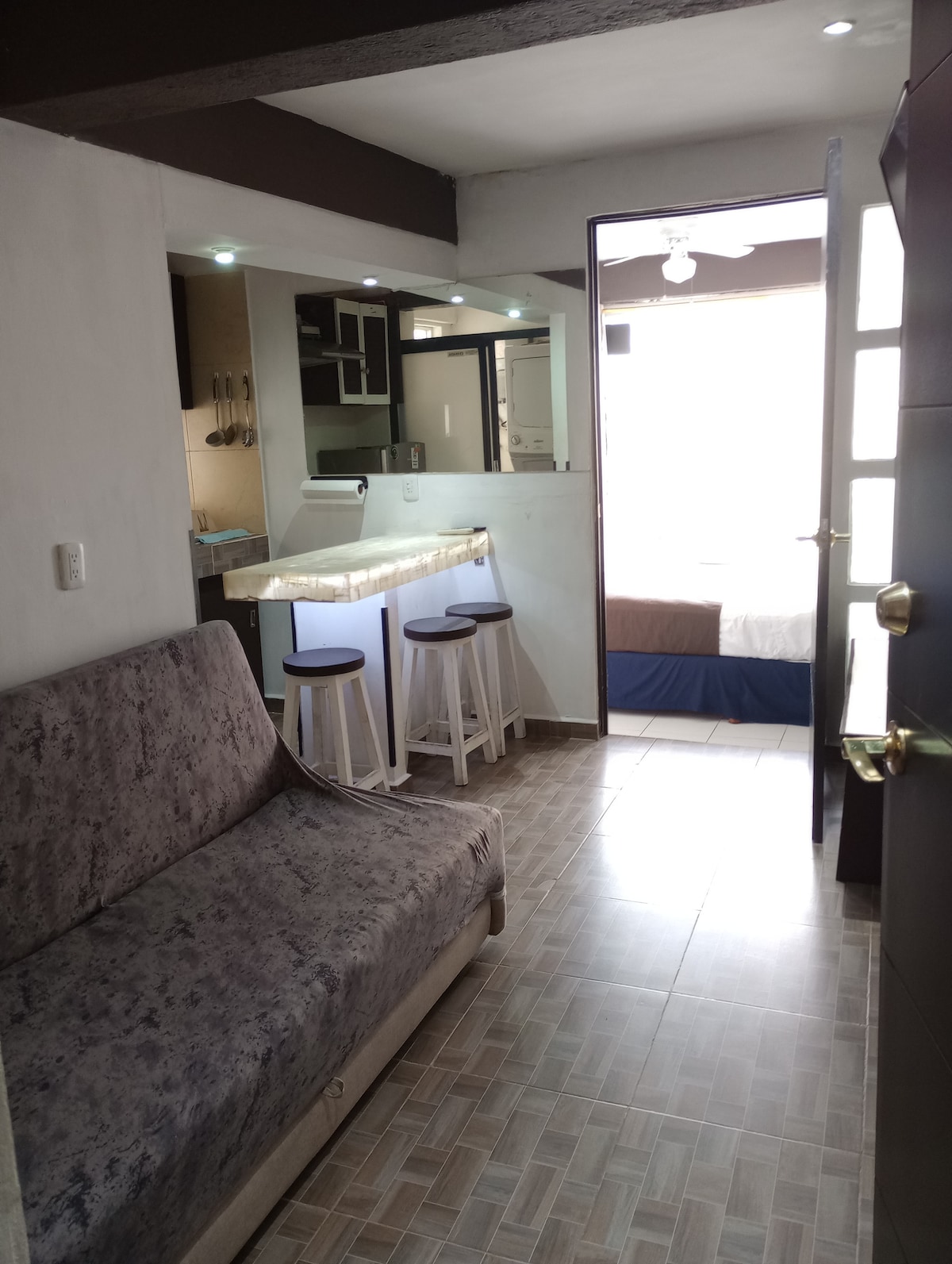 A small living area is visible, featuring a gray sofa and three white bar stools. The kitchenette and dining bar are located on the left, while a bright window illuminates the space. A bedroom can be seen in the background, with a bed framed by soft natural light.
