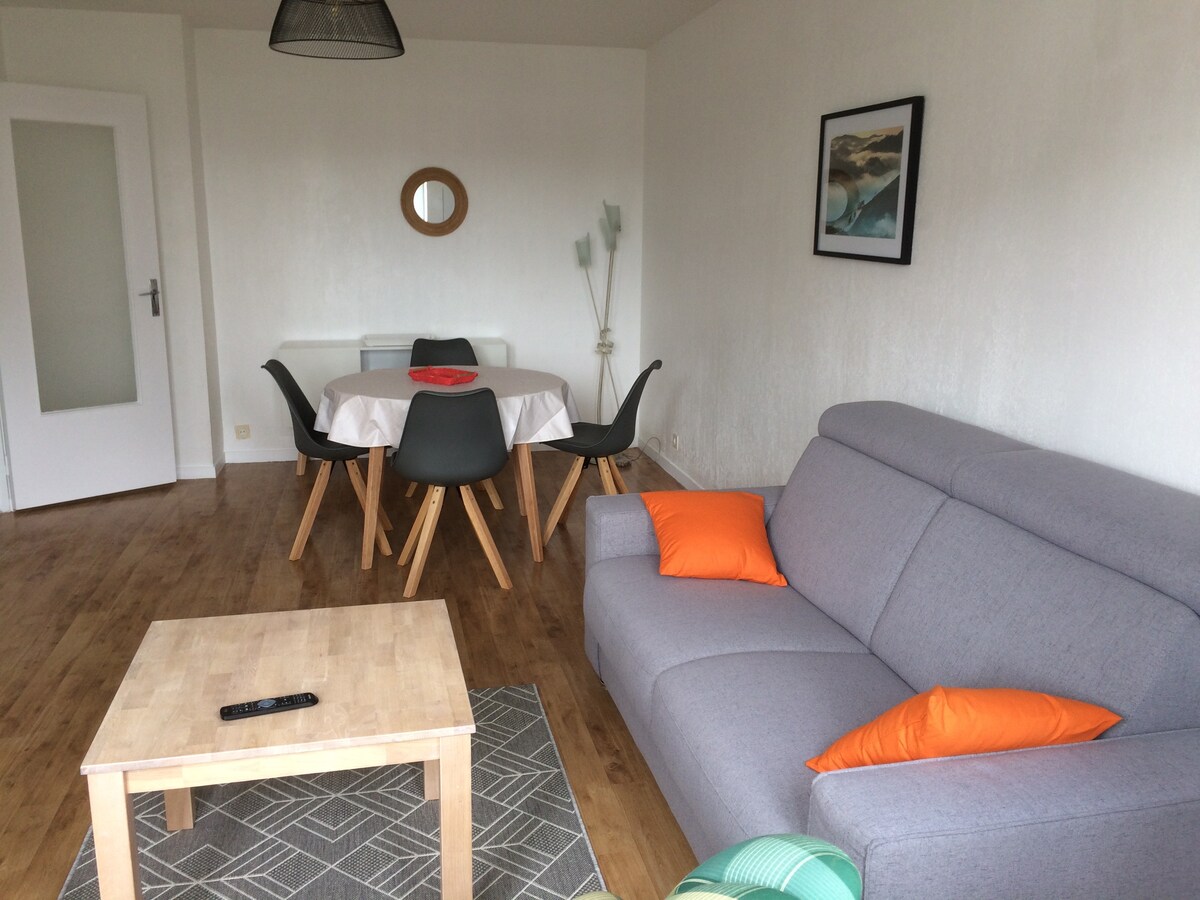 A bright living area features a gray sofa with orange cushions, complemented by a wooden coffee table. A round dining table with four chairs is positioned nearby. A wall-mounted artwork and a circular mirror add depth to the space. Natural light illuminates the room.