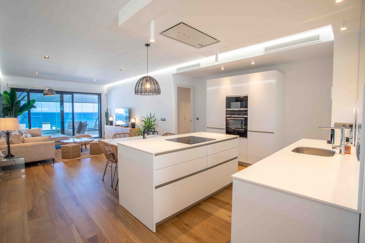 An open kitchen area features a modern island with a built-in sink and stove. Bright cabinetry lines the walls, while stylish pendant lights hang above. The space connects to a living area with large windows showcasing stunning sea views and natural light streaming in.