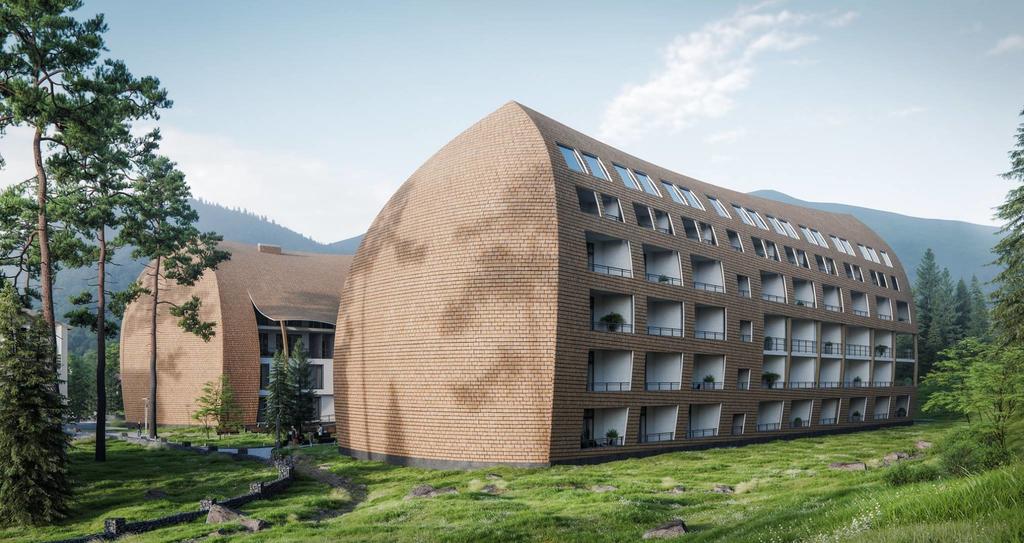 A modern multi-level building features an angular, textured exterior with large windows. Surrounding greenery includes trees and rocks, creating a peaceful natural setting. The building's design offers visual interest and blends with the scenic landscape of mountains and foliage.