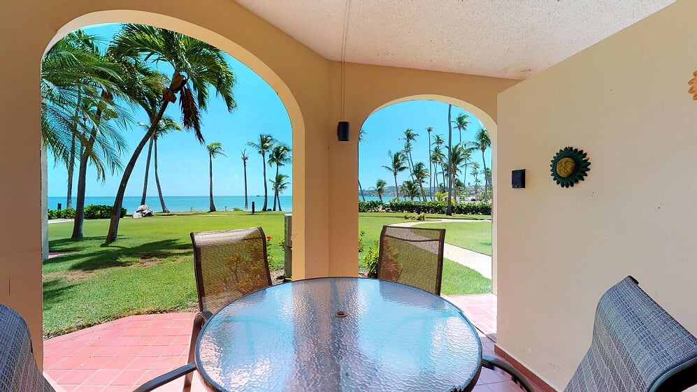 A covered outdoor patio is framed by archways, offering a view of palm trees and the ocean. A glass-top table is set with four metal chairs. Vibrant greens of the lawn and the distant shoreline create a serene backdrop.