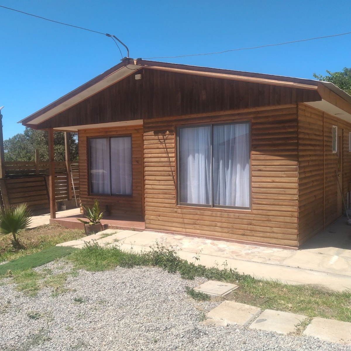 A wooden cabin stands under a clear blue sky, featuring large windows with sheer curtains that allow natural light to brighten the interior. A small garden area with gravel and stepping stones surrounds the entrance, enhancing the outdoor appeal.