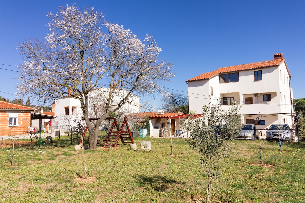 A spacious garden is showcased, featuring a large flowering tree and a playground swing set. In the background, two structures are visible, one with a terracotta roof and another with a light facade. The grass is well-maintained and surrounded by small plants.