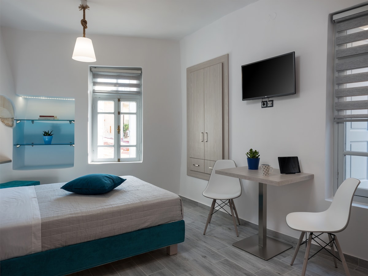 A bright and airy studio space features a spacious bed with a blue accent pillow. A small dining table with two chairs is positioned near a window, with natural light illuminating the area. A mounted television and a shelf with decorative plants add functionality.