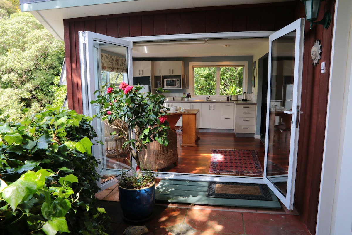 An open set of double doors reveals a bright kitchen area with modern appliances. A small dining table is visible, complemented by a large window showcasing views of greenery. Potted plants frame the entrance, enhancing the outdoor connection.