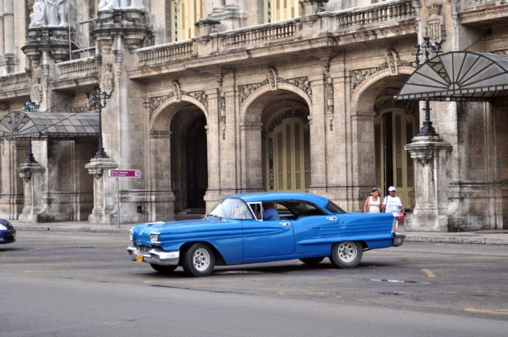 A vintage blue car is seen driving on a cobblestone street, with grand, historic buildings serving as a backdrop. The architecture features ornate details, large arched windows, and decorative elements that evoke the charm of Old Havana.