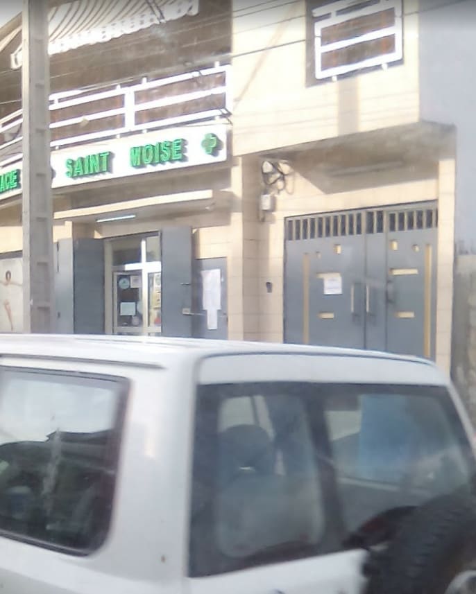 The exterior of the Saint Moïse pharmacy building is featured, showcasing green illuminated signage. Multiple large windows and a secure entry door are visible, framed by a neutral-colored facade. A white vehicle is parked nearby, blending into the streetscape.