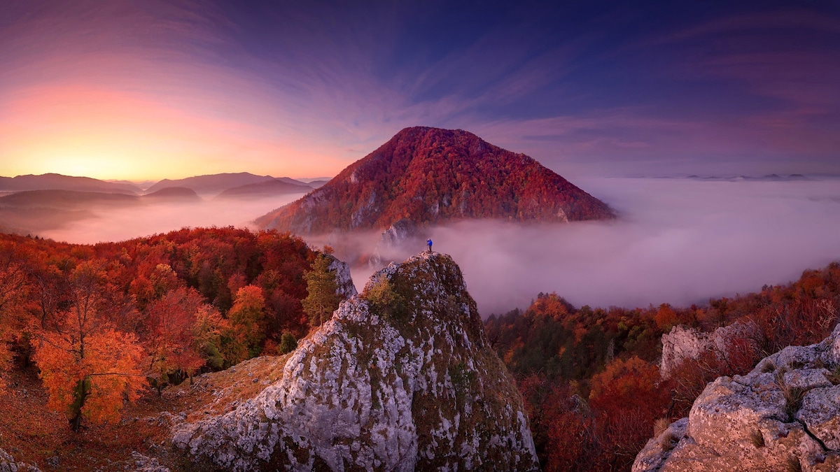 A panoramic view captures a vibrant autumn landscape with rolling hills and a foggy valley. The scene is dominated by a mountain bathed in warm light, while colorful foliage blankets the foreground. Soft wisps of fog create a serene atmosphere, enhancing the natural beauty of the region.