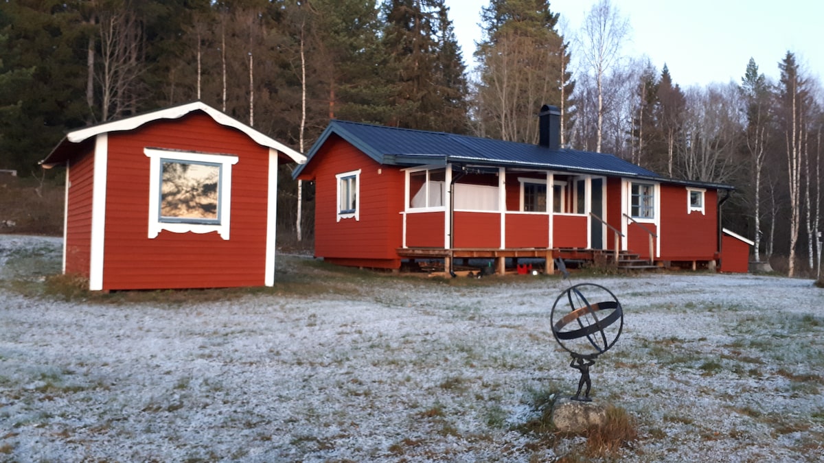 A charming red timber cabin is depicted surrounded by frost-covered grass. A smaller matching building stands nearby, featuring a window that reflects the serene landscape. A decorative sundial is located in the foreground, adding a rustic touch to the tranquil outdoor setting.