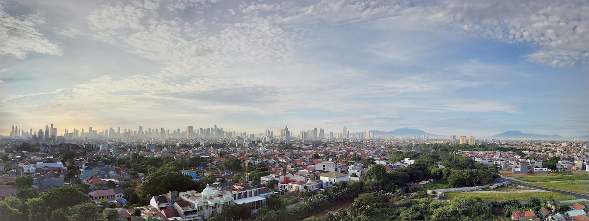 A panoramic view captures Jakarta's skyline, featuring a blend of skyscrapers and residential areas. The horizon showcases both urban development and green spaces, with distant mountains visible under a light blue sky adorned with clouds.