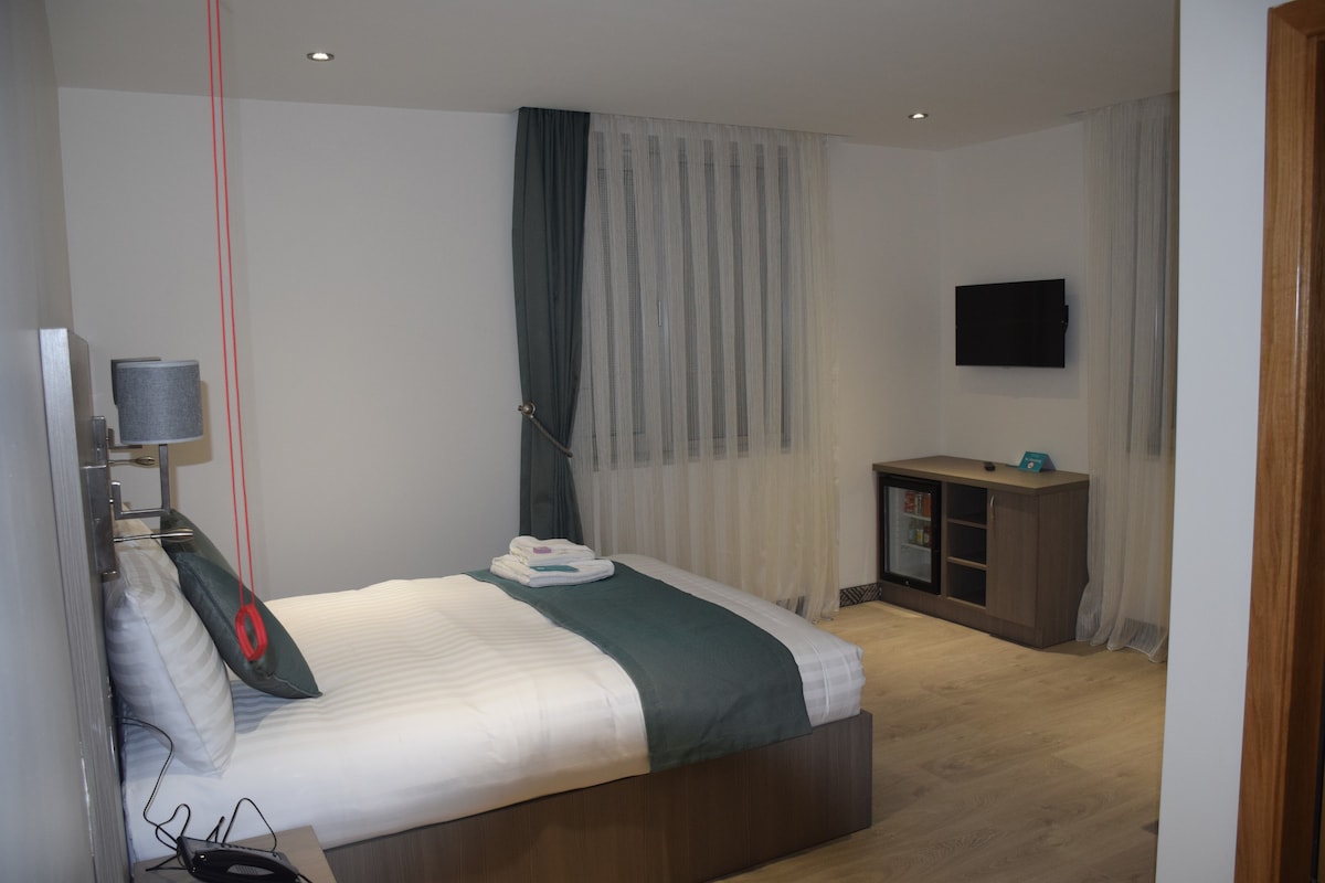 A modern room features a spacious bed with crisp linens and a decorative throw. Sheer curtains allow natural light to filter in. A 32" Smart TV is mounted on the wall across from a small cabinet, enhancing the minimalist design.