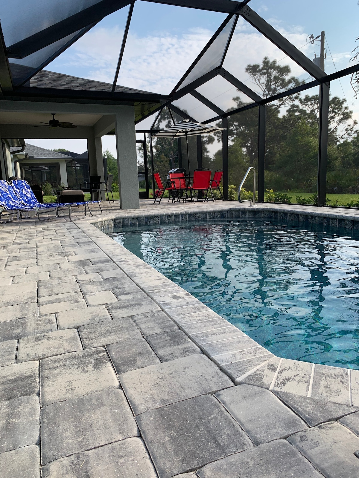 A spacious screened-in lanai surrounds a serene swimming pool. Comfortable lounge chairs and a dining set with red chairs are visible near the pool. The patio features textured stone flooring, and lush greenery can be seen beyond the screen.