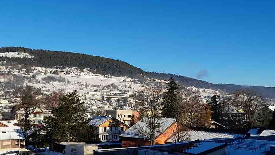 A scenic view captures snow-covered hills and a serene landscape. The foreground features trees and buildings with a backdrop of a clear blue sky, creating an inviting atmosphere. The hillside is blanketed in white snow, hinting at the winter season.