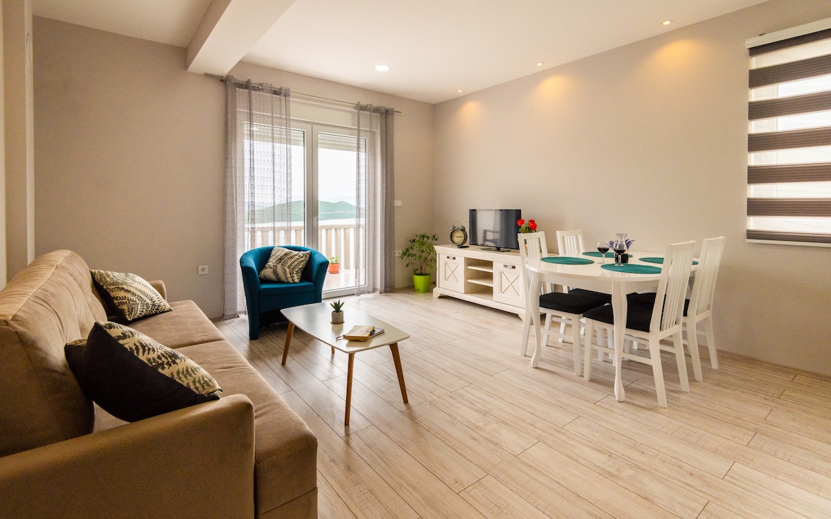 A modern living area features a light-colored sofa and a turquoise accent chair. A dining table is surrounded by white chairs, positioned beside a large window that provides natural light. A flat-screen TV is situated on a white cabinet, enhancing the inviting space.