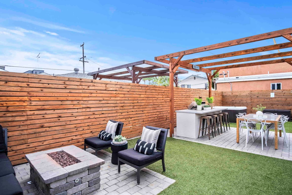 An outdoor retreat features a comfortable seating area with two lounge chairs surrounding a stone fire pit. A dining area with a long table and chairs is positioned beside a modern outdoor kitchen. Wooden fencing and greenery add privacy to the space.