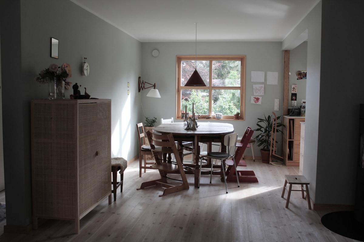A dining area is featured, showcasing a round table surrounded by various wooden chairs. Natural light filters through a large window, highlighting the gentle hues of the walls and the greenery outside. A sideboard and plants add to the welcoming environment.