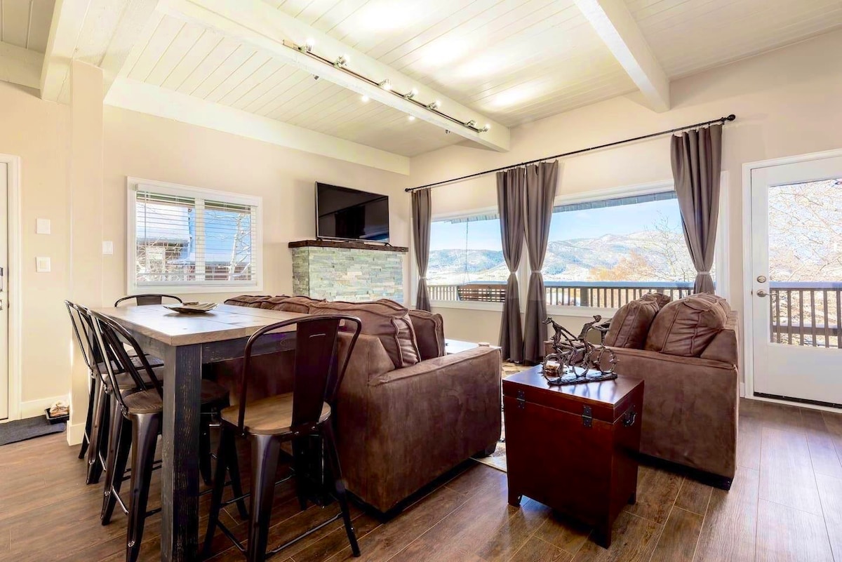 A cozy living area features a comfortable seating arrangement with two brown sofas and a coffee table. Large windows provide expansive views of the surrounding landscape, while a flat-screen TV is mounted above a stone fireplace. Natural light brightens the space, which is decorated with soft curtains.