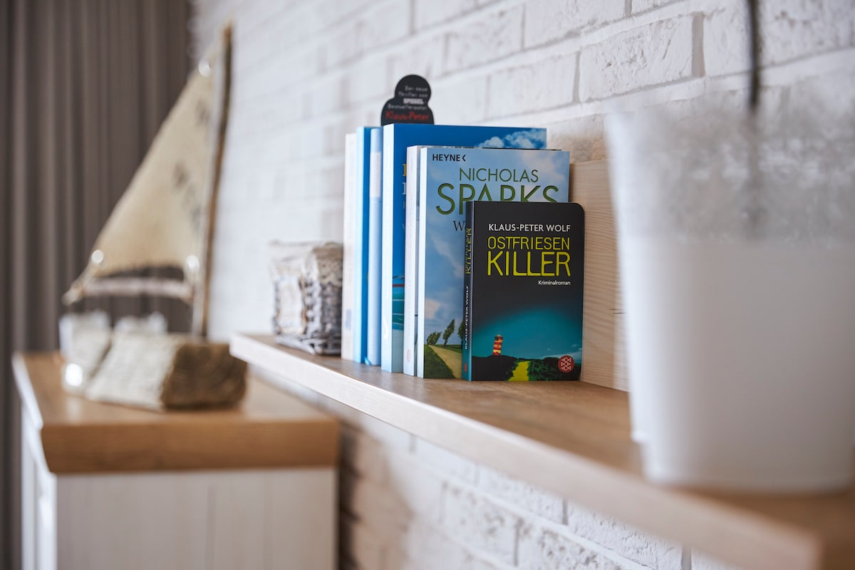 A wooden shelf displays a selection of books, including titles by Nicholas Sparks and Klaus-Peter Wolf. A decorative item resembling a sailboat can be seen in the background, alongside neatly stacked towels placed on the shelf.
