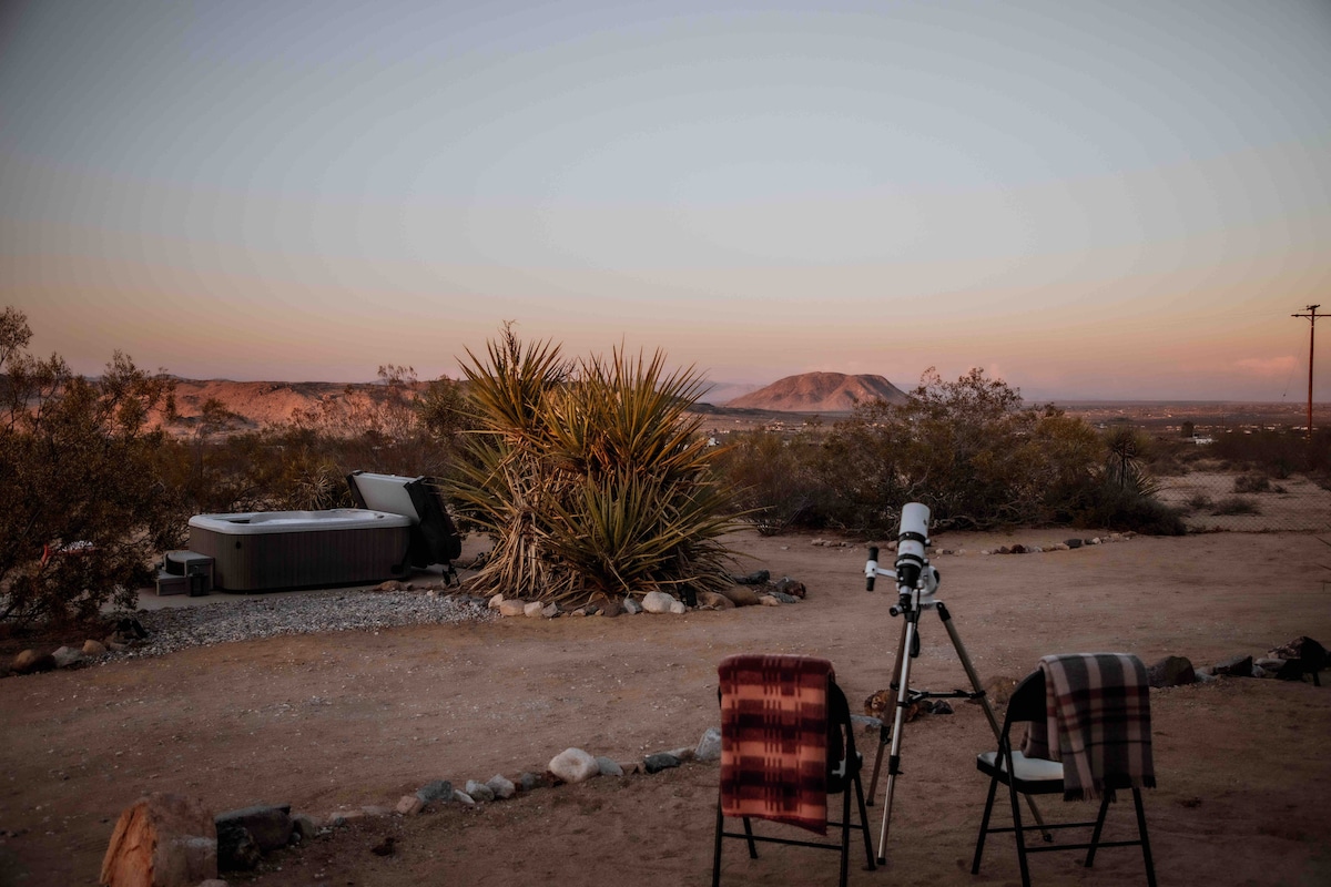 A telescope is positioned on a stable tripod, facing the expansive desert landscape under a fading twilight sky. Two chairs are placed nearby, one covered with a blanket. The hot tub is visible in the background, surrounded by desert foliage and hills.