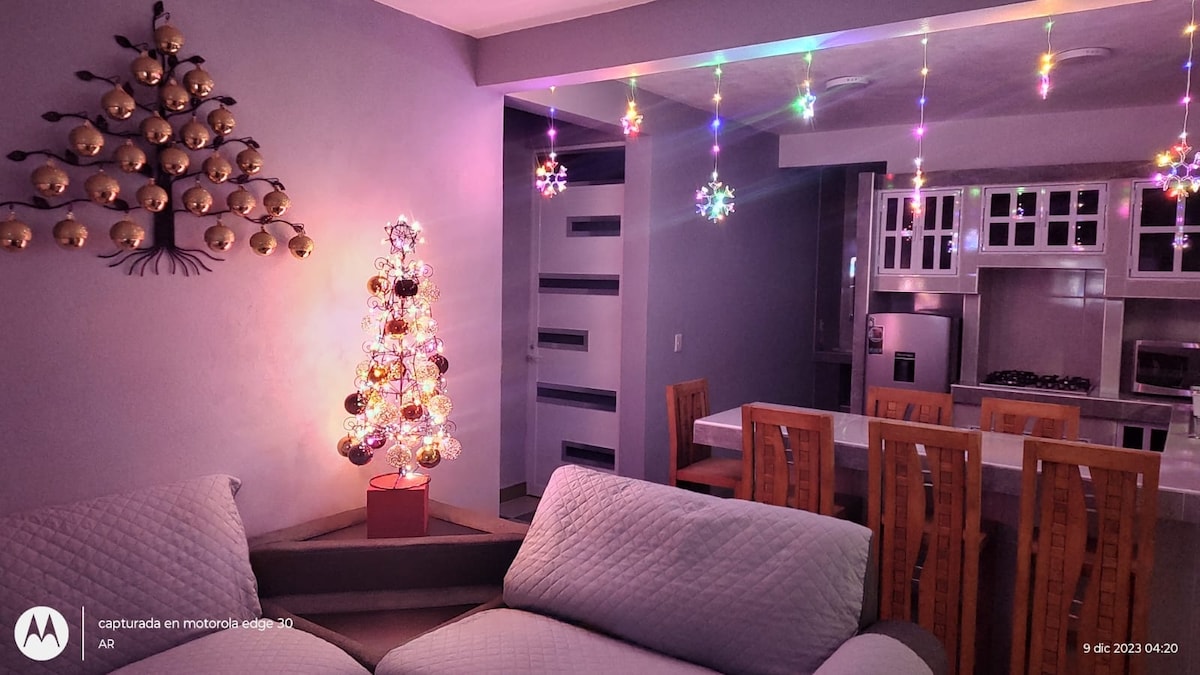A cozy living area is highlighted by a colorful, decorated Christmas tree beside a modern couch. String lights hang from the ceiling, creating a festive ambiance. A dining table with six wooden chairs is visible in the background, accompanied by an open kitchen space.