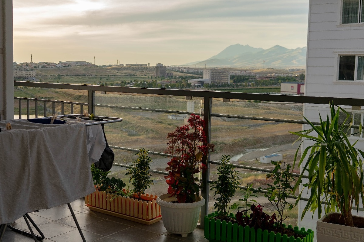 A balcony features a scenic view of rolling hills and distant mountains. Potted plants and decorative gardening borders line the balcony, adding touches of greenery. The space includes a clothes drying rack, suggesting a functional outdoor area for relaxation.