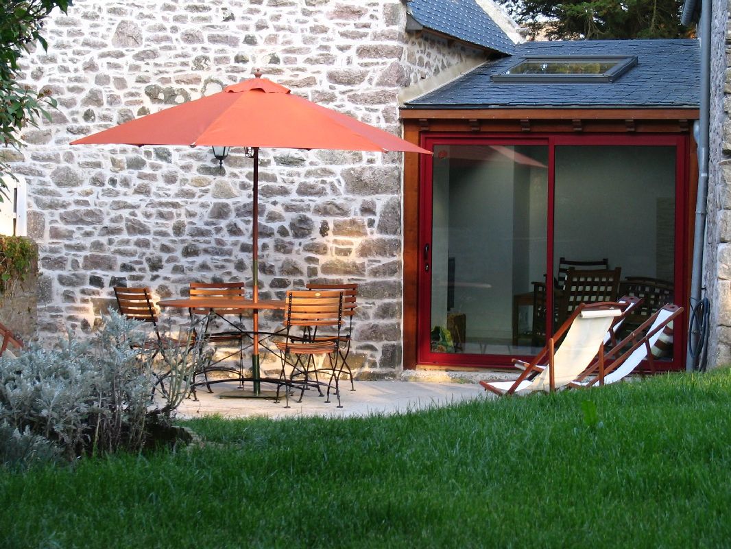A patio area is shown featuring a round wooden table surrounded by several metal chairs, shaded by a large orange umbrella. Adjacent to the patio, sliding glass doors provide access to a bright indoor space. A grassy area with low shrubbery enhances the outdoor setting.